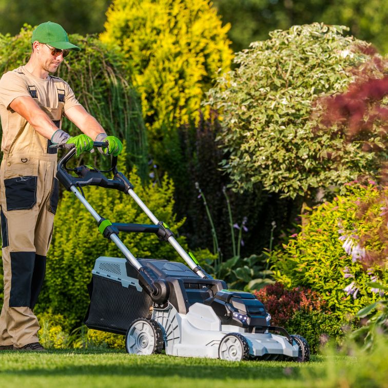 Worker maintaining a large estate garden by trimming hedges, mowing the lawn, and planting flowers near elegant buildings.